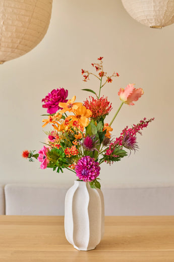 In een witte keramische vaas staat het boeket ‘Magnus di Lambo’ van Zijden kunstbloemen, een kleurrijk arrangement van roze, oranje en gele kunstbloemen met groene bladeren, geplaatst op een houten tafel tegen een beige achtergrond.