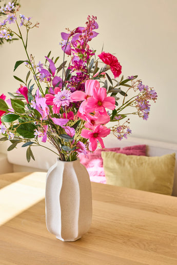 In een witte keramische vaas staat het boeket ‘Purple Dawn’ van Zijden kunstbloemen, een levendig arrangement van roze, paarse en magenta bloemen; de vaas staat op een lichte houten tafel met kleurrijke kussens op de achtergrond.