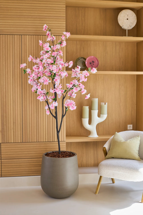 Un fiore di ciliegio artificiale rosa di 180 cm, un albero in vaso con fiori rosa, si trova accanto a una sedia bianca con cuscini in una stanza moderna con scaffali in legno e oggetti decorativi.