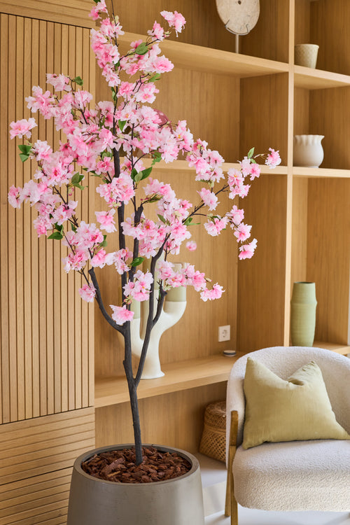 Il fiore di ciliegio artificiale rosa da 150 cm, con i suoi delicati fiori di seta rosa, sembra elegante in una stanza moderna con scaffali in legno, oggetti decorativi, una sedia beige e un cuscino verde - aggiungendo il fascino dei fiori di seta ai tuoi interni.