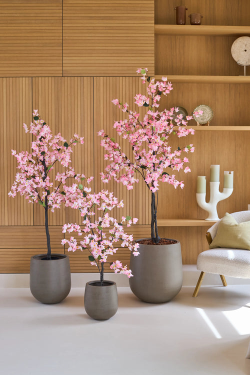 Tre piante in vaso del tipo Bougainvillea artificiale da 150 cm, di colore rosa con fiori rosa, sono disposte davanti a ripiani in legno, accanto a una moderna sedia bianca con cuscino e un candeliere decorativo.