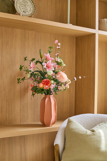 Su uno scaffale di legno accanto a un'accogliente sedia con un cuscino verde c'è un vaso rosa con il bouquet di fiori artificiali di seta "Yasmine's Choice", che crea un'immagine interna calda e invitante.