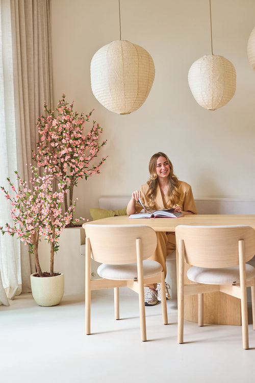 Una donna con lunghi capelli biondi è seduta a un tavolo di legno chiaro, legge un libro e sorride. Accanto a lei c'è la pianta artificiale di fiori di ciliegio rosa da 150 cm, i cui fiori rosa forniscono un accento vivace. Le lampade a sospensione e l'arredamento minimalista creano un'atmosfera leggera e accogliente.
