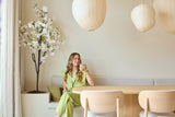 Een vrouw in een lichtgroen pak zit op een bankje aan een houten eettafel, met een kopje in haar hand. Boven haar hangen decoratieve papieren lantaarns en in de hoek staat de Kunst Kersenbloesem 180 cm wit, terwijl er zacht natuurlijk licht door een raam naar binnen valt.