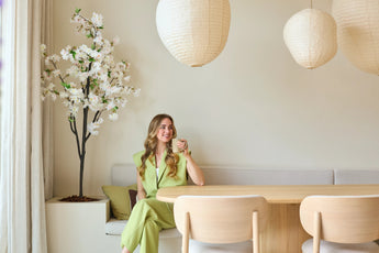 Een vrouw in een lichtgroen pak zit op een bankje aan een houten eettafel, met een kopje in haar hand. Boven haar hangen decoratieve papieren lantaarns en in de hoek staat de Kunst Kersenbloesem 180 cm wit, terwijl er zacht natuurlijk licht door een raam naar binnen valt.