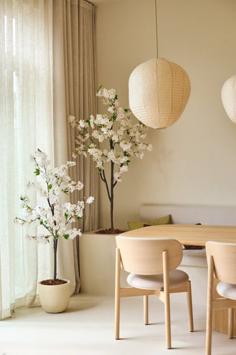 Een minimalistische eethoek met stoelen van licht hout, een ronde tafel, twee grote papieren lantaarns, doorschijnende gordijnen en kunstplanten van Kunst Kersenbloesem van 180 cm met witte kunstmatige kersenbloesembomen, die een rustige en luchtige sfeer creëren.