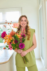 Een glimlachende vrouw met lang blond haar, gekleed in een lichtgroene outfit, staat binnenshuis met in haar handen het Zijden kunstbloemenboeket Magnus di Lambo — een kleurrijk, met de hand gebonden boeket van zijden bloemen, perfect voor wie de voorkeur geeft aan een onderhoudsvrij bloemenarrangement.