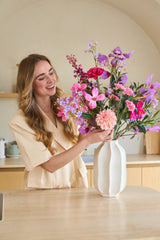 Een glimlachende vrouw met lang blond haar zet een kleurrijk boeket ‘Purple Dawn’ van Zijden kunstbloemen in een witte vaas op het aanrecht.