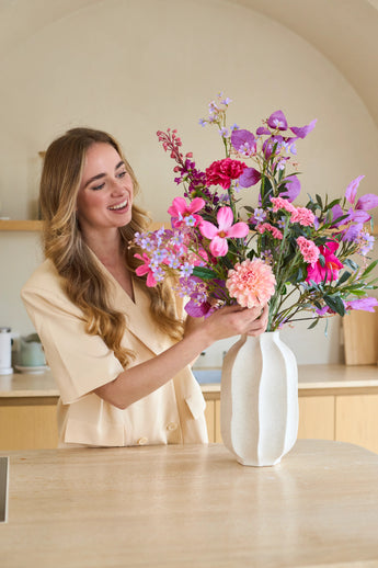 Een glimlachende vrouw met lang blond haar zet een kleurrijk boeket ‘Purple Dawn’ van Zijden kunstbloemen in een witte vaas op het aanrecht.