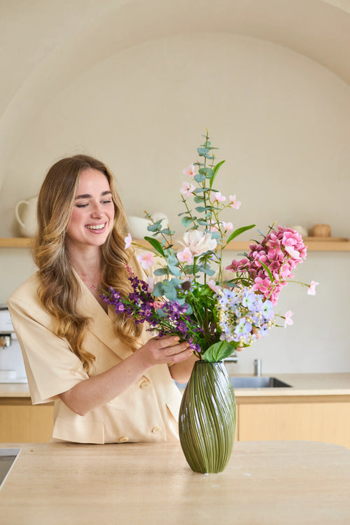 Una donna dai capelli lunghi e ondulati sistema un bouquet di fiori di seta artificiale "Lavender Dreams" in un vaso verde nella sua cucina moderna, sorridendo mentre lavora. Il vibrante bouquet di fiori di seta porta una bellezza senza tempo nella sua casa.