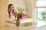 Una donna sorridente che sistema il bouquet "Fiori di seta artificiale Svenski colorati" in un vaso verde su un piano di lavoro chiaro in una cucina con arredamento moderno e luce naturale.