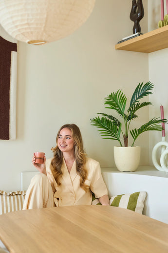 Una donna sorridente con lunghi capelli biondi siede in una stanza moderna e luminosa con una tazza rosa in mano. Intorno a lei ci sono piante verdi, tra cui la rigogliosa pianta della Fenice Artificiale alta 85 cm, oltre a mensole in legno e oggetti decorativi.
