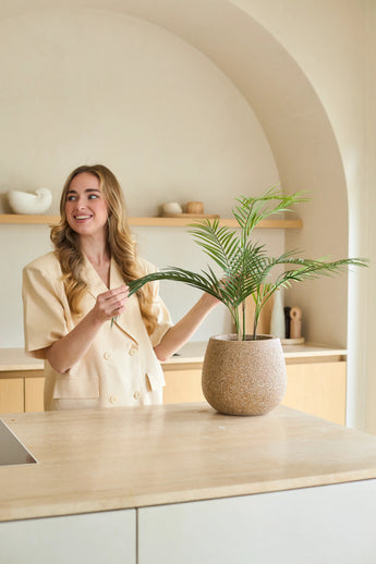 Una donna con lunghi capelli biondi sorride mentre si prende cura di una palma artificiale Chamaedorea in vaso da 18 pollici in una cucina moderna dai toni neutri con un arredamento minimalista.
