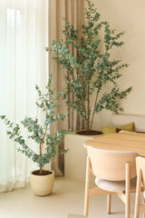 Due piante artificiali di eucalipto artificiale di 150 cm, posizionate accanto a tende trasparenti e pareti beige, abbinate a un tavolo da pranzo e sedie in legno chiaro in un interno minimalista e moderno.