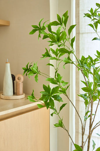 Una pianta artificiale Pieris Japonica di 150 cm si trova vicino a una finestra accanto a un mobile in legno chiaro, che contiene bottiglie di ceramica e un macinapepe, aggiungendo un tocco minimalista alla stanza luminosa e ariosa.
