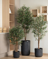 Een 160 cm grote Kunst olijfboom met groene bladeren staat in een zwarte geribbelde plantenbak op een lichthouten vloer naast beige kasten en open wandplanken.