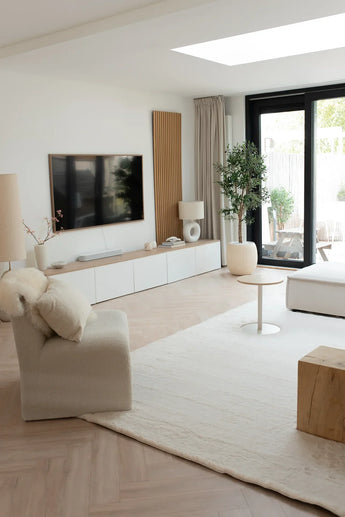 Een moderne, minimale woonkamer in neutrale tinten heeft een pluche stoel, een wit vloerkleed, een houten salontafel, een tv aan de muur en een Kunst olijfboom van 160 cm UV. Grote glazen deuren leiden naar een patio met buitenzitjes en de ruimte wordt gevuld met natuurlijk licht.