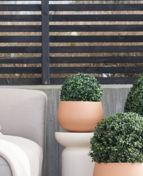 Tre piante artificiali topiarie rotonde di bosso stile romano UV D30 cm si trovano su un patio vicino a una sedia da esterno beige, contro una moderna recinzione a doghe nere e alberi spogli.