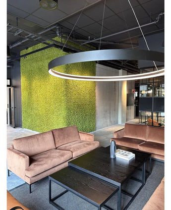 Una moderna area salotto con divani beige, tavolini neri, una grande lampada da soffitto rotonda e una parete verde sullo sfondo, realizzata con Kunsthaag Nordic Moss 50x50 cm UV, che irradia interni lussuosi e un design industriale contemporaneo.