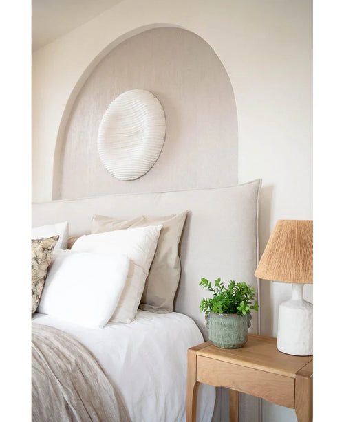 Un'accogliente camera da letto con testiera beige, biancheria da letto e cuscini neutri, un comodino in legno sormontato da una pianta di eucalipto artificiale da 27 cm, una lampada con paralume strutturato e decorazioni murali bianche rotonde sopra il letto in un'alcova a volta.