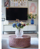 Een moderne woonkamer heeft een terrazzo salontafel met een glazen vaas met bloemen, een tv aan de muur, ingelijste kunst en foto's, een wit dressoir en de Kunst Hortensia Deluxe 57 cm lila als elegant blauw bloemstuk op de achtergrond.