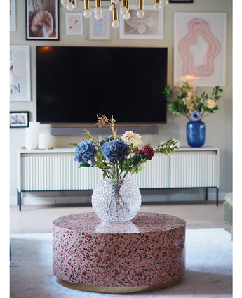 Een moderne woonkamer heeft een terrazzo salontafel met een glazen vaas met bloemen, een tv aan de muur, ingelijste kunst en foto's, een wit dressoir en de Kunst Hortensia Deluxe 57 cm lila als elegant blauw bloemstuk op de achtergrond.