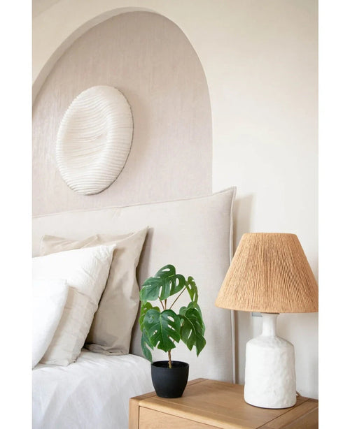 Un letto ben fatto con biancheria bianca si trova accanto a un comodino in legno con l'Art Monstera 40 cm in un vaso decorativo nero e una lampada bianca con paralume intrecciato. Alla parete sono appesi un arco morbido e un'opera d'arte rotonda e strutturata.