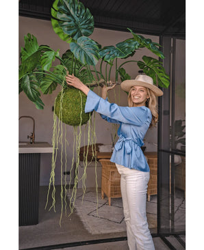 Una donna sorridente con una camicetta blu, pantaloni bianchi e cappello mostra la pianta pensile artificiale Monstera Deluxe 80 cm con sinuosi tralci verdi, che dona un'atmosfera tropicale al luminoso spazio interno con pareti di vetro e mobili in rattan.