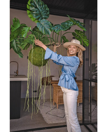 Een glimlachende vrouw in een blauwe blouse, witte broek en hoed toont de Kunst hangplant Monstera Deluxe 80 cm met slingerende groene wijnranken, die een tropische sfeer brengt in de lichte binnenruimte met glazen wanden en rotan meubilair.