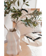 Een wit gebeeldhouwde vaas houdt Kunst olijfblad 90 cm UV met groene bladeren op een lichte houten tafel. Daarachter versterken een beige stoel en twee crèmekleurige kussens met opvallende zwarte strepen de mediterrane sfeer.