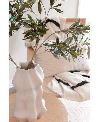 Een wit gebeeldhouwde vaas houdt Kunst olijfblad 90 cm UV met groene bladeren op een lichte houten tafel. Daarachter versterken een beige stoel en twee crèmekleurige kussens met opvallende zwarte strepen de mediterrane sfeer.