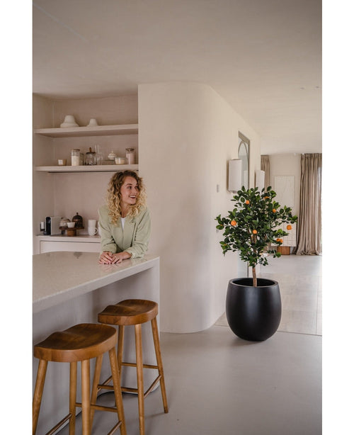 Una donna dai capelli biondi e ricci sorride dietro un bancone con due sgabelli di legno. L'Arancio artificiale da 120 cm in vaso aggiunge fascino allo spazio moderno e minimalista decorato con tonalità neutre.