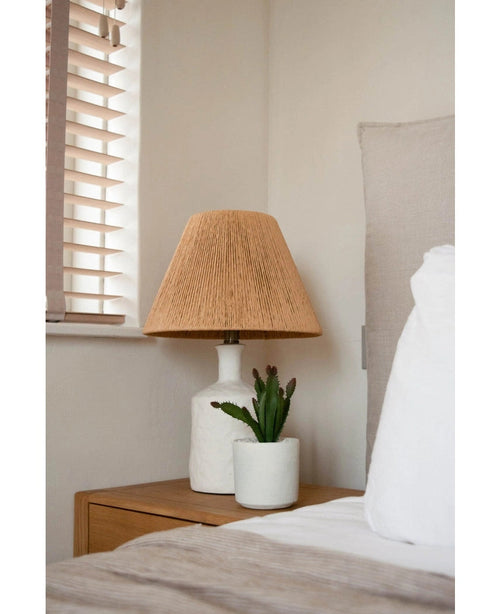 Un comodino con una lampada bianca con paralume intrecciato beige e una pianta grassa artificiale di 20 cm si trova accanto a un letto con lenzuola bianche e beige, vicino a una finestra con persiane in legno.