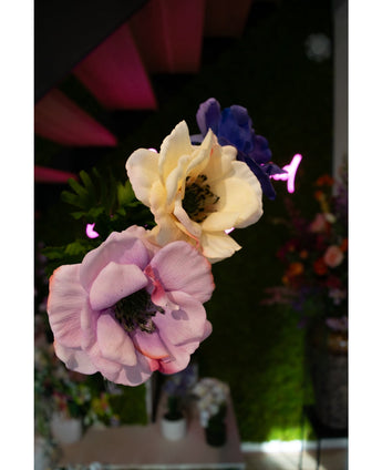 Close-up van drie Kunstanemoon Real Touch geel 52cm kunstanemoonbloemen - lavendel, crème en blauw - tegen vaag groen gebladerte en neonroze verlichting, met bloemstukken in de verte in beeld.