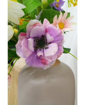Een close-up van de Kunstanemoon Real Touch paars 52cm anemoon bloem in een mat witte vaas, omringd door groene bladeren en pastelkleurige bloemen.