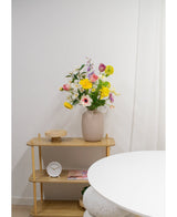 Een beige vaas met Kunstanemoon Real Touch paars 52cm bloemen staat op een licht houten plank naast een kleine ronde klok en een dienblad. Op de voorgrond staat een witte ronde tafel tegen een witte muur en gordijn.