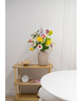Een beige vaas met Kunstanemoon Real Touch paars 52cm bloemen staat op een licht houten plank naast een kleine ronde klok en een dienblad. Op de voorgrond staat een witte ronde tafel tegen een witte muur en gordijn.