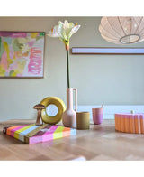 Een moderne houten tafel toont kleurrijke objecten, waaronder een roze vaas met daarin de Kunstbloem Amaryllis Real Touch wit 66 cm, een gele ringsculptuur, gestreepte boeken en een roze beker. Abstracte kunst accentueert de achtergrondmuur.