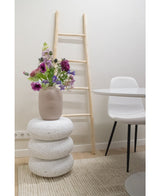 Een Kunstbloem Grasklokje 62 cm paars staat in een beige vaas op gestapelde witte terrazzo krukjes. Daarachter staat een lichthouten ladder, bij een witte stoel en een ronde tafel, allemaal geplaatst op een beige vloerkleed met textuur in een minimalistische kamer.