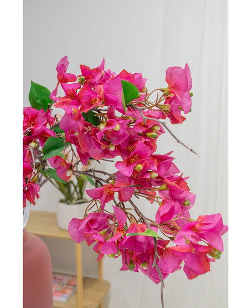 Un primo piano del fiore artificiale Bougainvillea viola da 90 cm mostra i suoi vivaci fiori viola e le foglie verdi all'interno, con una pianta in vaso su una mensola di legno e un muro bianco leggermente sfocato sullo sfondo.