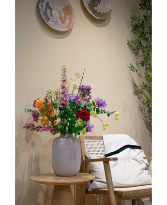 Een witte vaas met Kunstkornoelje Real Touch Lichtgeel 74cm staat op een houten bijzettafeltje naast een lichthouten stoel met een wit kussen en een kleed met zwarte randen, geaccentueerd door geweven wanddecoratie en een groene plant op de achtergrond.