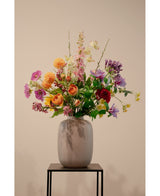 De Kunstkornoelje Real Touch Lichtgeel 74cm, een levensechte lichtgele kunstbloem, staat in een keramieken vaas met groen gebladerte op een zwart metalen standaard tegen een effen beige achtergrond.