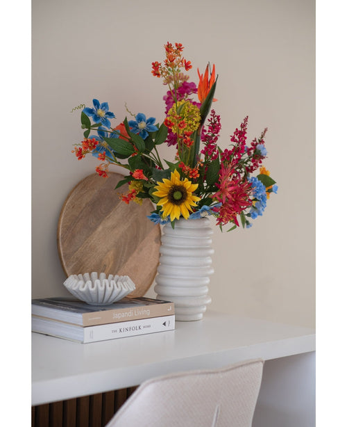 Un mazzo di fiori di garofano artificiale di 70 cm di colore blu in un vaso di ceramica bianca si trova su una scrivania bianca accanto a libri impilati e una piccola ciotola decorativa, con un vassoio rotondo di legno appoggiato al muro dietro di esso.