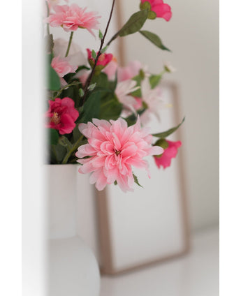 Een close-up van de Kunstbloem Dahlia 68 cm roze in een witte vaas, met een vaag fotolijstje op de achtergrond op een wit vlak.