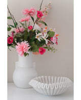 Een Kunstbloem Dahlia 68 cm roze in een witte vaas staat naast een schaal van wit keramiek met ruches op een wit oppervlak, tegen een effen lichte achtergrond.