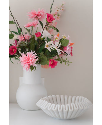 Een Kunstbloem Dahlia 68 cm roze in een witte vaas staat naast een schaal van wit keramiek met ruches op een wit oppervlak, tegen een effen lichte achtergrond.