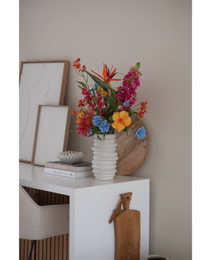Un vaso bianco a coste con fiore artificiale di erba gatta rosa da 75 cm si trova su una scrivania bianca con libri, opere d'arte incorniciate, un vassoio di legno e un tagliere, accanto a una sedia beige in una stanza moderna e minimalista.