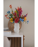 Un vaso bianco strutturato contiene un bouquet di fiori artificiali di erba gatta rosa da 75 cm su un tavolo bianco accanto a libri e decorazioni impilati, con un vassoio rotondo di legno appoggiato al muro beige sullo sfondo.