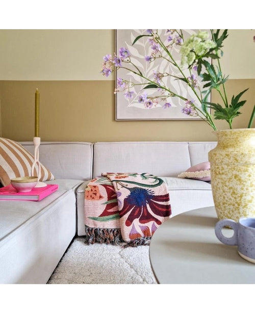 Un accogliente soggiorno con un divano beige chiaro, un plaid floreale, una candela rosa, un cuscino a righe e un fiore artificiale in pizzo bianco da 76 cm in un vaso giallo sul tavolo rotondo. L'arte astratta decora il muro dietro di esso.
