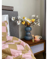 Een nachtkastje met een blauwe vaas met daarin de Kunstbloem Magnolia 50 cm geel/oranje, een roze theekopje op een margrietvormig schoteltje en een brandende kaars staat naast een bed dat is bedekt met een roze en bruine deken met geometrische patronen.